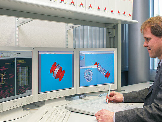 An engineer reviews 3D CAD models of a red mechanical component on dual computer monitors while taking notes at a desk.