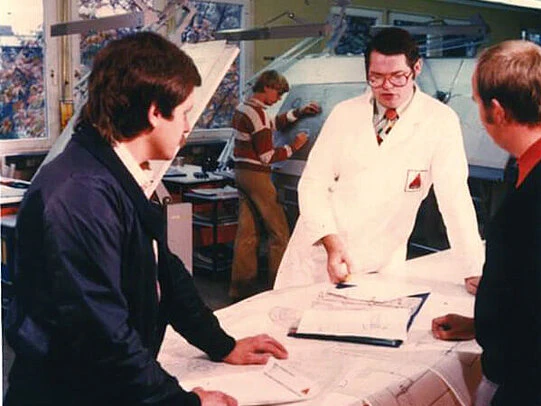 Engineer in a white lab coat reviews technical drawings with colleagues around a drafting table in a design office.