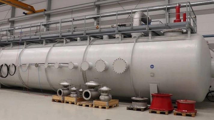 Large horizontal industrial pressure vessel inside a factory hall, equipped with multiple flanged nozzles, valves on pallets, and an elevated service platform.