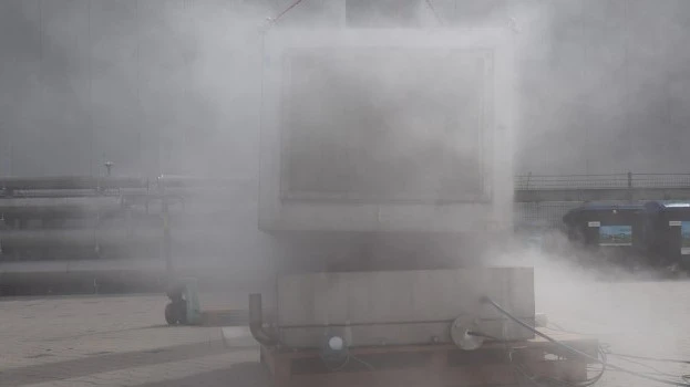 Outdoor industrial test setup with a large container largely obscured by a dense cloud of venting vapor or smoke during an active test.
