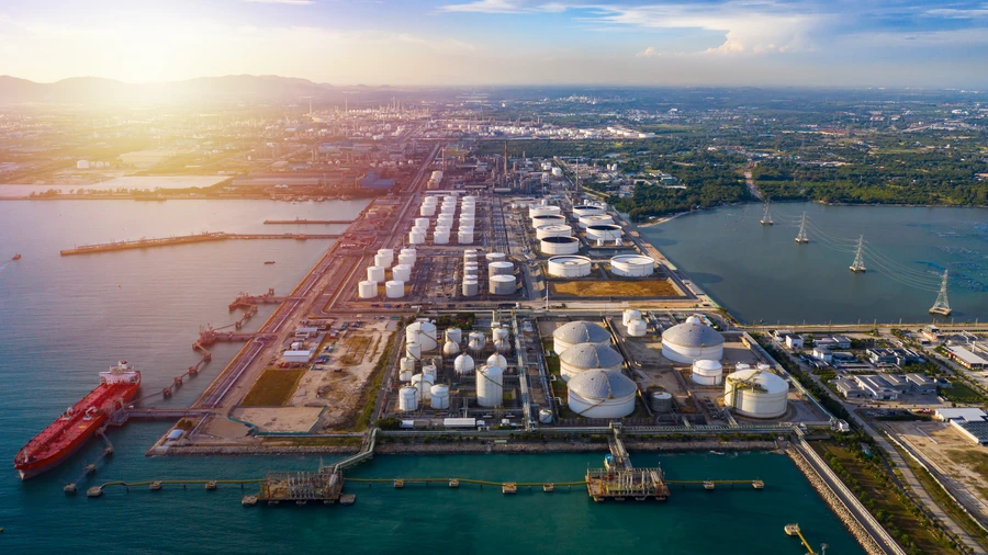 Aerial view of a coastal industrial refinery with rows of white storage tanks, docks with a red tanker ship, and waterways stretching into the distance at sunset.