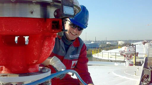 A worker in red coveralls walks through a large industrial plant filled with pipes, valves, and tall processing towers.