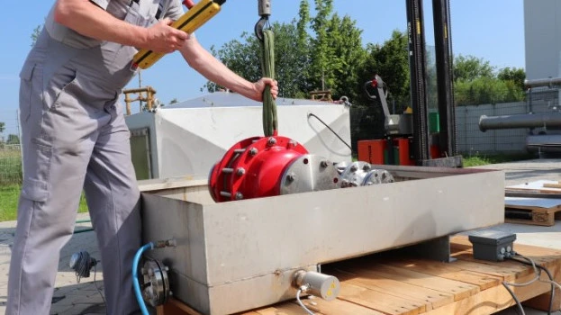 A technician lowers a large red industrial safety device into a rectangular outdoor test basin using a hoist, with cables and piping connected.