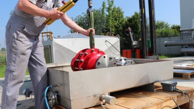 A technician lowers a large red industrial safety device into a rectangular outdoor test basin using a hoist, with cables and piping connected.