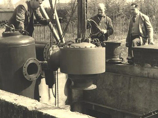 Vintage black-and-white photo of three men installing or inspecting a large industrial valve assembly over a concrete pit outdoors.