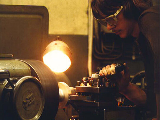 Person wearing protective goggles operates a metal lathe in a workshop, illuminated by a bright task lamp.