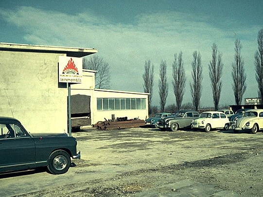 Vintage photo of a light-colored industrial building with a “Leinemann & Co.” flame logo sign mounted on the wall, several 1950s-era cars parked in a gravel lot, and tall leafless trees lined up in the background.
