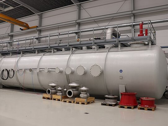 Large horizontal industrial pressure vessel inside a factory hall, equipped with multiple flanged nozzles, valves on pallets, and an elevated service platform.