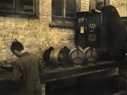 Vintage workshop scene showing workers beside cylindrical metal components arranged on a bench against a brick wall in an industrial setting.