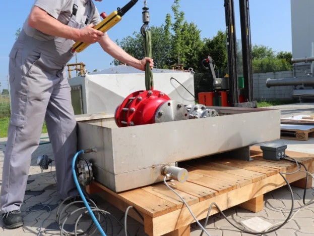 A technician lowers a large red industrial safety device into a rectangular outdoor test basin using a hoist, with cables and piping connected.