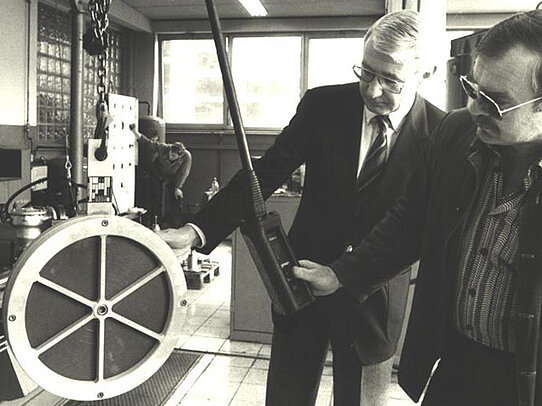 Two men in suits examine a large circular industrial component in a workshop, one holding a handheld measuring device while a worker stands in the background.