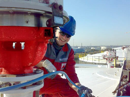A worker in red coveralls walks through a large industrial plant filled with pipes, valves, and tall processing towers.