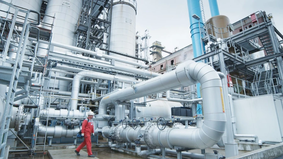 A worker in red coveralls walks through a large industrial facility filled with interconnected pipes, valves, and tall processing towers.