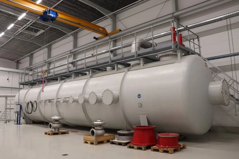 A large horizontal industrial pressure vessel sits inside a factory hall, with a platform and overhead crane above it.