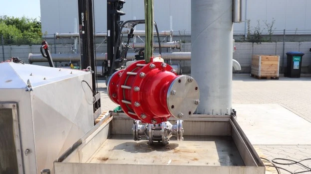 A large red industrial inline safety device is suspended by lifting straps above a metal test basin at an outdoor facility, with piping and equipment nearby.