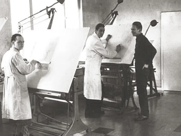 Three men in a black-and-white photo stand at drafting tables in a workshop, working on large technical drawings under adjustable lamps.