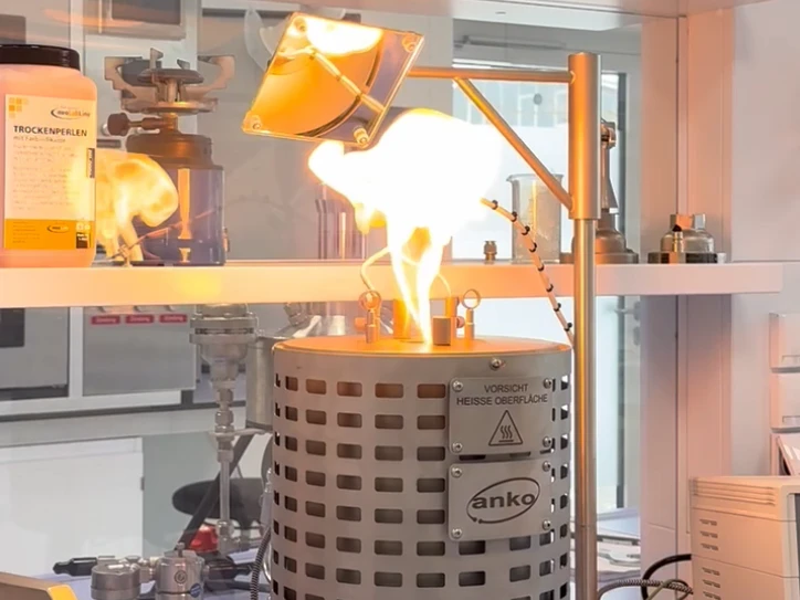 A laboratory burner flares with a bright yellow flame above a metal heating vessel under a mounted heat lamp.