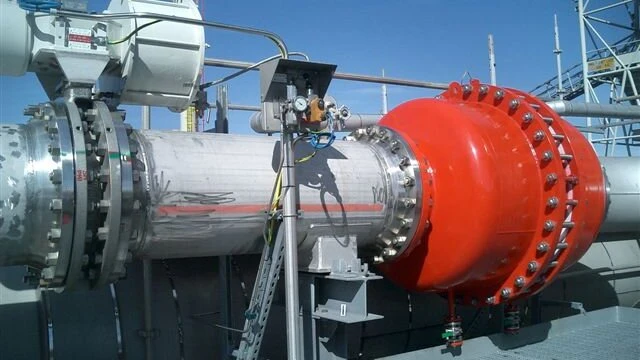 Close-up of an industrial pipeline with a large red flanged housing attached to a shiny metal pipe outdoors at a plant site.