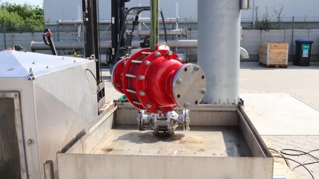 A large red industrial inline safety device is suspended by lifting straps above a metal test basin at an outdoor facility, with piping and equipment nearby.