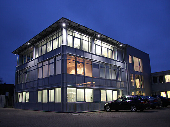 Modern glass-fronted office building illuminated at night with parked cars outside.