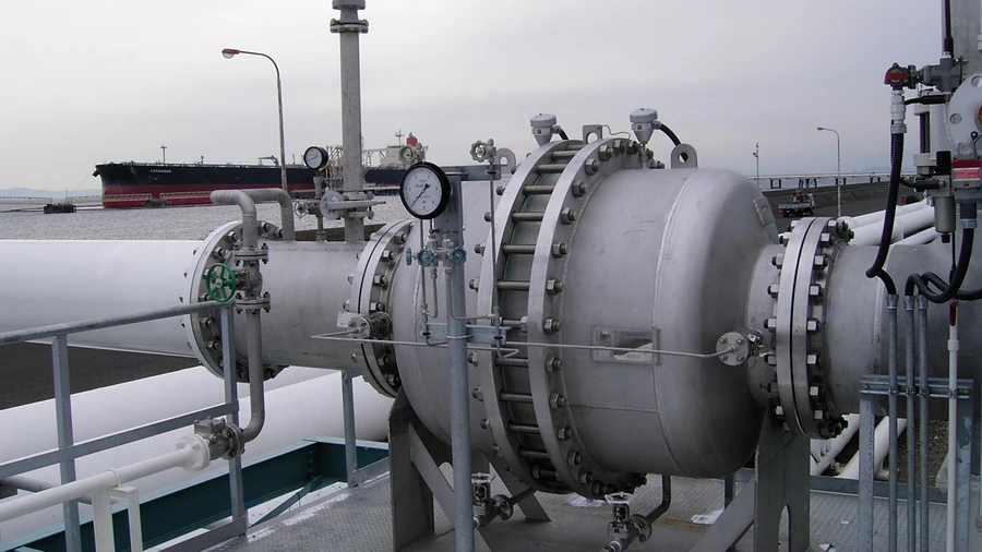 Eine Industriepipeline mit einem großen, verschraubten Druckbehälter und Messgeräten steht auf einer Plattform am Wasser, im Hintergrund ist ein Frachtschiff zu sehen.