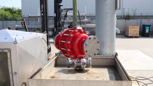 A large red industrial inline safety device is suspended by lifting straps above a metal test basin at an outdoor facility, with piping and equipment nearby.
