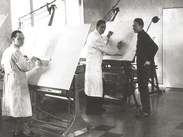 Three men in a black-and-white photo stand at drafting tables in a workshop, working on large technical drawings under adjustable lamps.