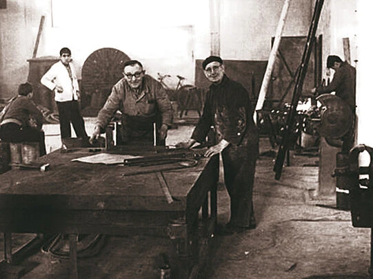 Black-and-white photo of several workshop workers gathered around a large worktable, reviewing metal parts and tools in an industrial manufacturing space.