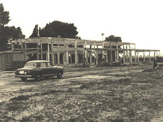 Vintage black-and-white photo of a car parked beside a partially constructed industrial building with exposed concrete framework.