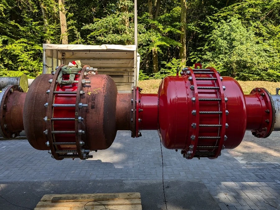 Side-by-side comparison of two large flanged industrial pipeline devices outdoors, one heavily corroded and one newly painted red, mounted between steel pipes against a wooded background.