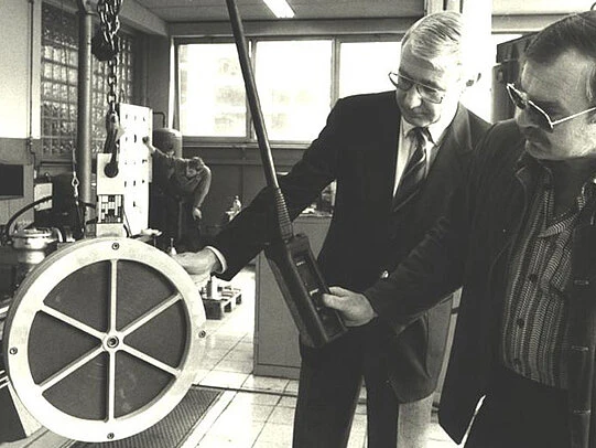 Two men in suits examine a large circular industrial component in a workshop, one holding a handheld measuring device while a worker stands in the background.