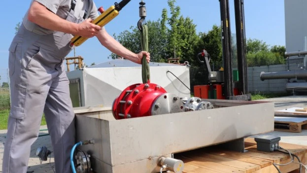 A technician lowers a large red industrial safety device into a rectangular outdoor test basin using a hoist, with cables and piping connected.