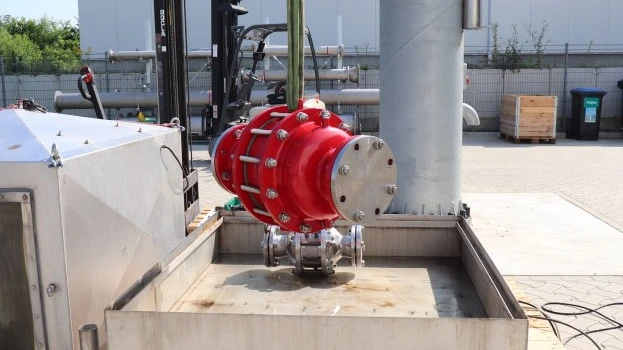 A large red industrial inline safety device is suspended by lifting straps above a metal test basin at an outdoor facility, with piping and equipment nearby.