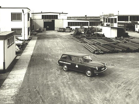 Vintage black-and-white photo of an industrial yard with factory buildings, stacked metal pipes, and a company car parked in the center.