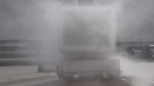 Outdoor industrial test setup with a large container largely obscured by a dense cloud of venting vapor or smoke during an active test.