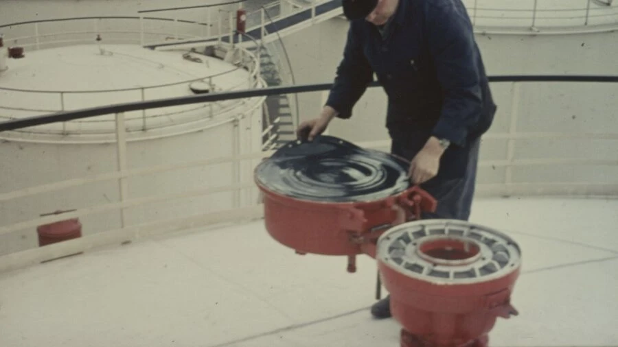 A worker on top of a large storage tank lifts the lid of a red circular hatch or vent.