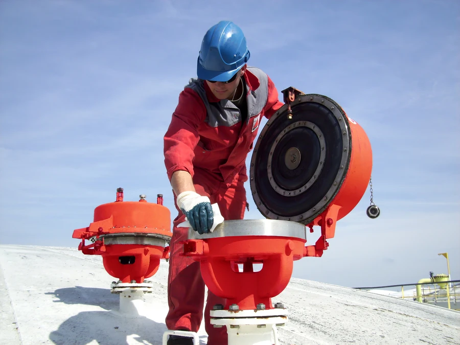 A worker in red coveralls and a blue hard hat opens and inspects a large red vent valve on top of a storage tank under a clear sky.