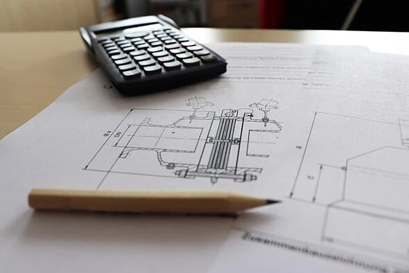 Close-up of an engineering drawing on paper with a pencil and calculator resting on top, suggesting technical design or calculation work.