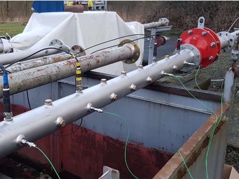 Outdoor industrial test setup with a horizontal pipe over an open containment pit, featuring a red flanged device and multiple sensors connected by green cables.