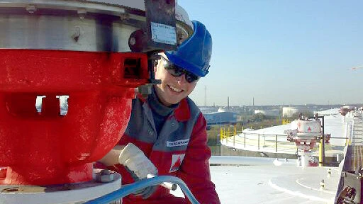 A worker in red coveralls walks through a large industrial plant filled with pipes, valves, and tall processing towers.