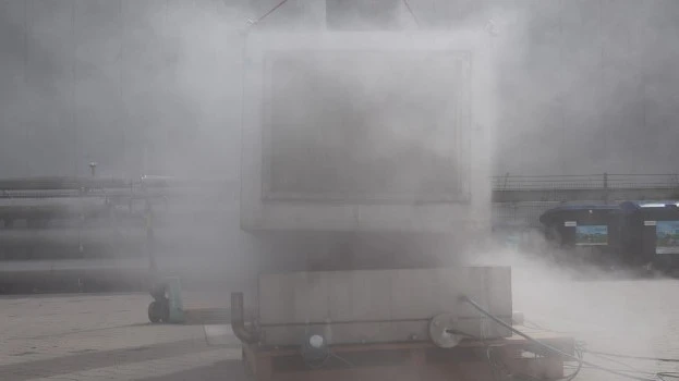 Outdoor industrial test setup with a large container largely obscured by a dense cloud of venting vapor or smoke during an active test.