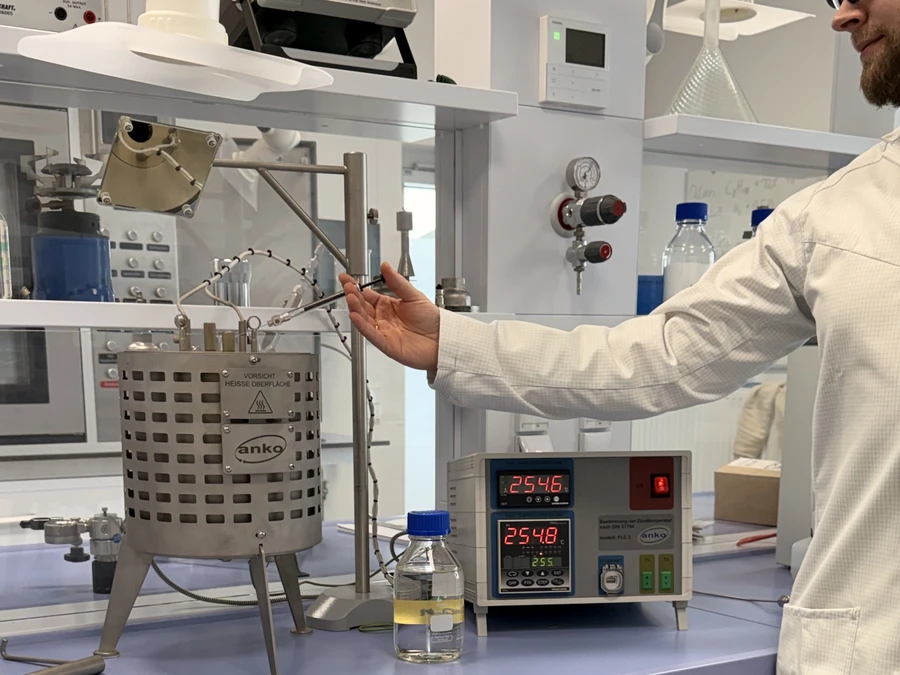A person in a lab coat operates a heated metal vessel in a laboratory, with a digital control unit and bottles on the bench.