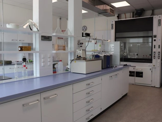 A clean laboratory workbench with cabinets, glassware, and a large fume hood in the background.