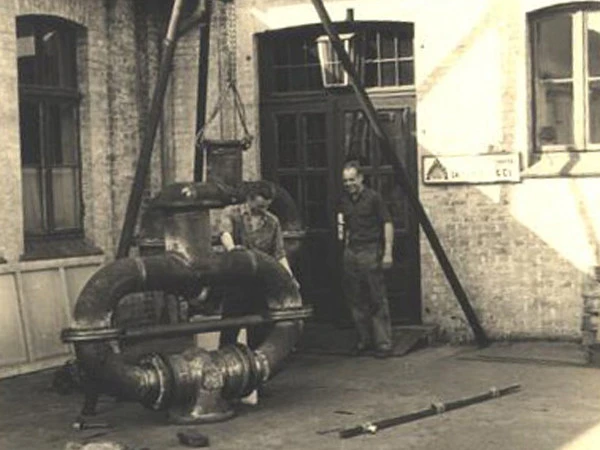 Vintage black-and-white photo of two men standing beside a large industrial pipe assembly being lifted in a brick courtyard.