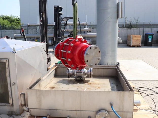 A large red industrial inline safety device is suspended by lifting straps above a metal test basin at an outdoor facility, with piping and equipment nearby.
