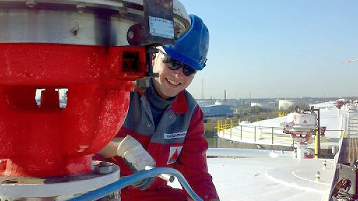 A worker in red coveralls walks through a large industrial plant filled with pipes, valves, and tall processing towers.