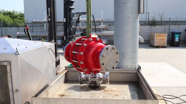 A large red industrial inline safety device is suspended by lifting straps above a metal test basin at an outdoor facility, with piping and equipment nearby.