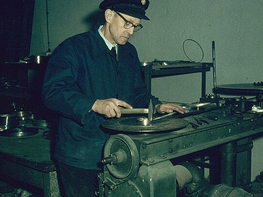 A technician in a workshop carefully measures a circular metal component on a workbench using hand tools and gauges.