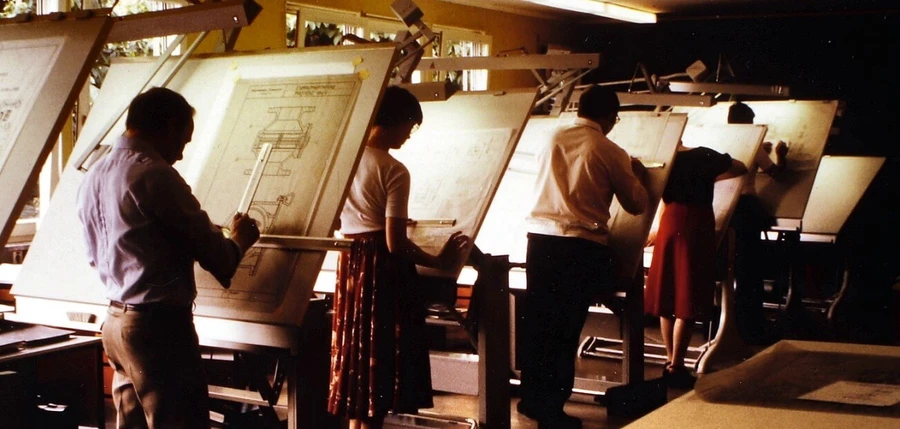 Several people stand at angled drafting tables in a vintage design studio, working on large technical drawings under warm indoor lighting.