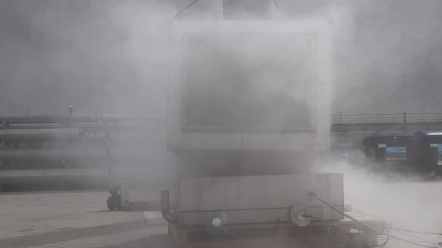 Outdoor industrial test setup with a large container largely obscured by a dense cloud of venting vapor or smoke during an active test.